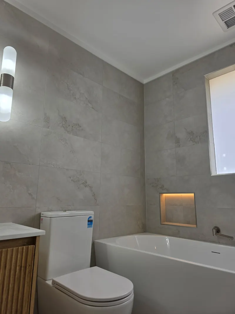 Finished bathroom with a built in bath under a lit tiled niche, beside a toilet and a wall light.
