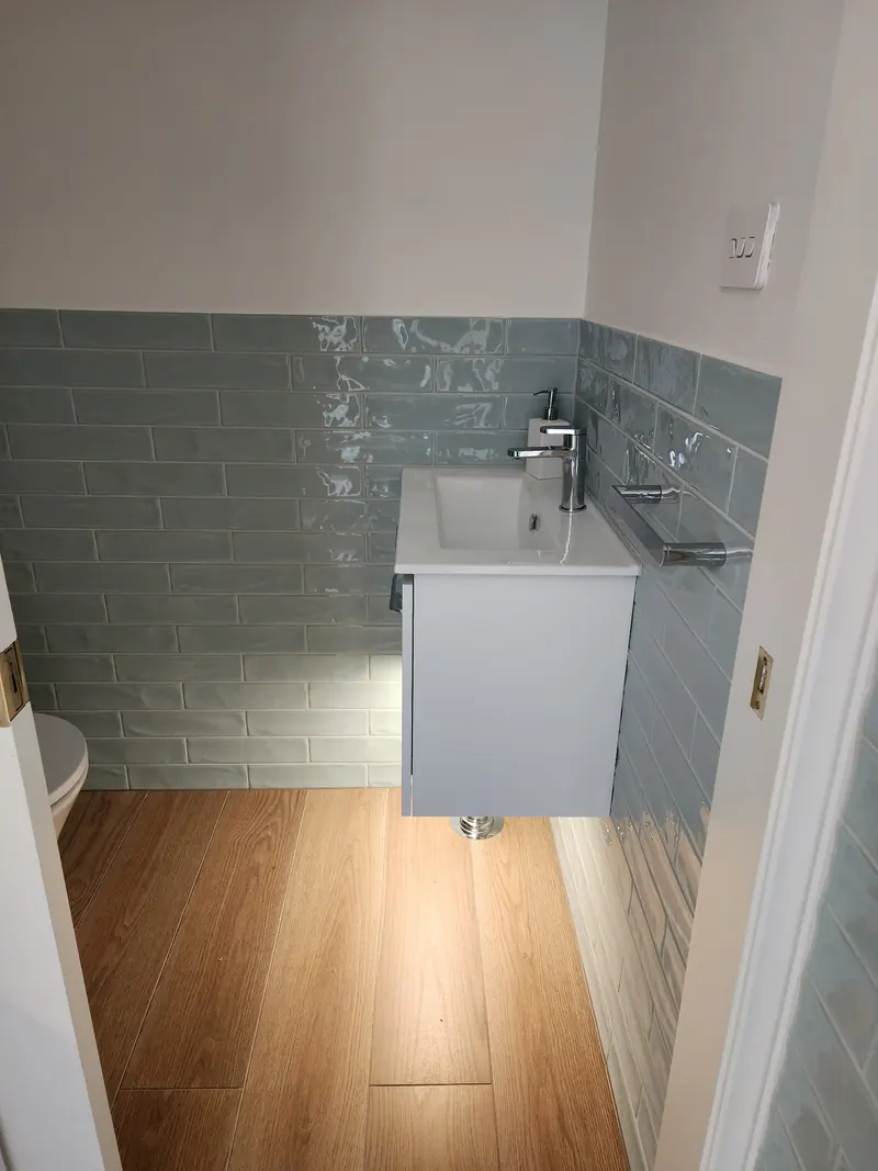 Small wall hung vanity and mixer against a green subway tiled wall above a timber look floor.