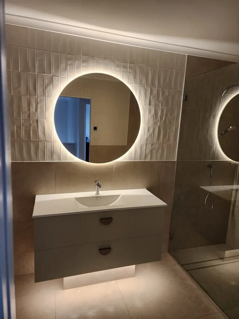 Finished bathroom with a wall hung vanity, a backlit round mirror and a walk in shower alongside.