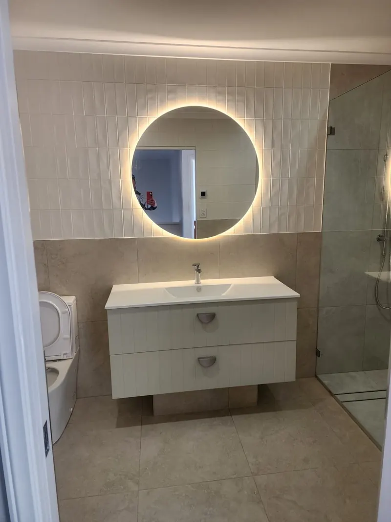 Finished bathroom with a white wall hung vanity under a backlit round mirror, and the shower screen to one side.