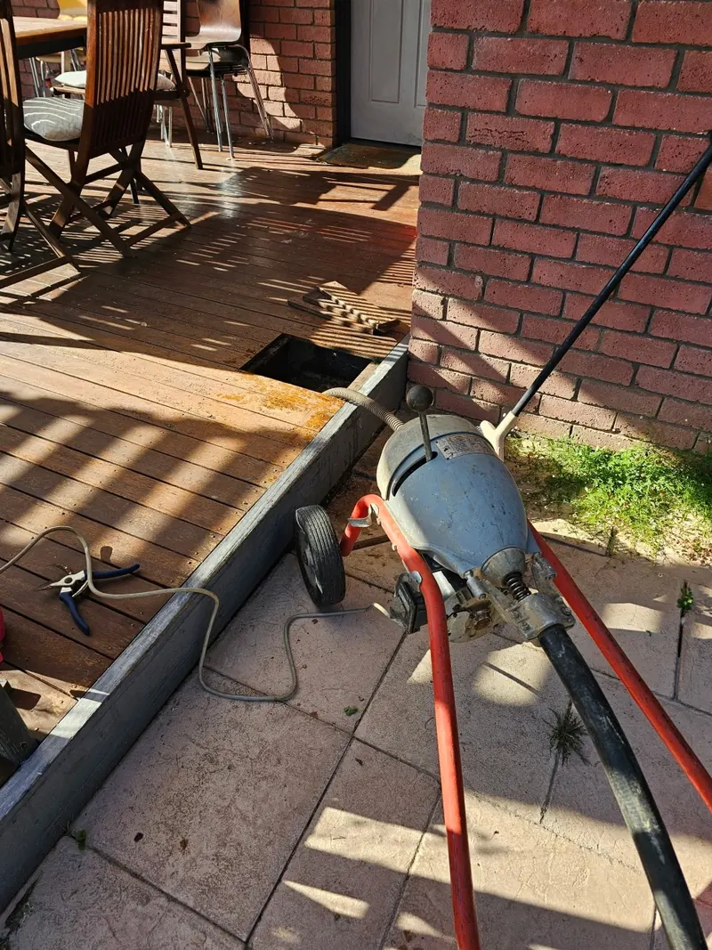 A drain cleaning machine and hose run out to a lifted drain cover at the edge of a paved courtyard.