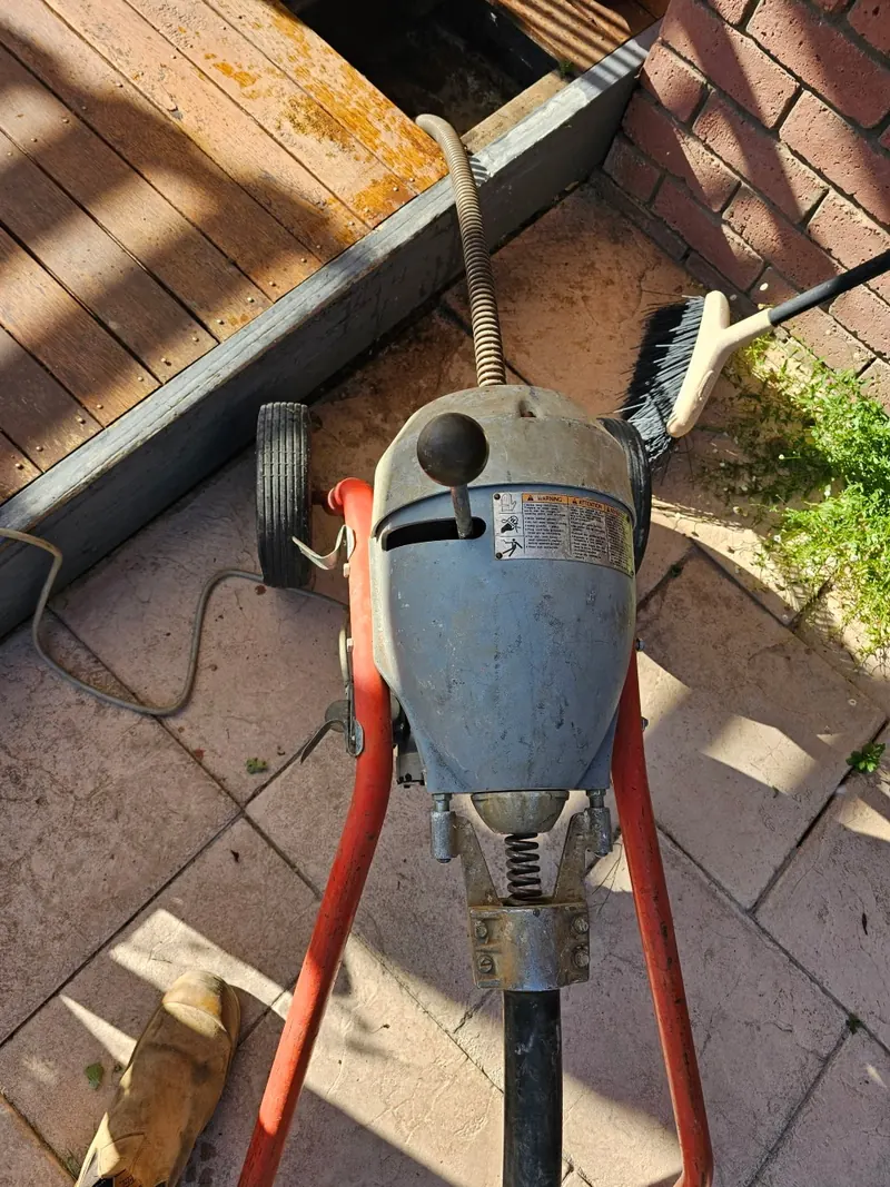 A drain cleaning machine set up over a lifted drain cover beside a timber deck.