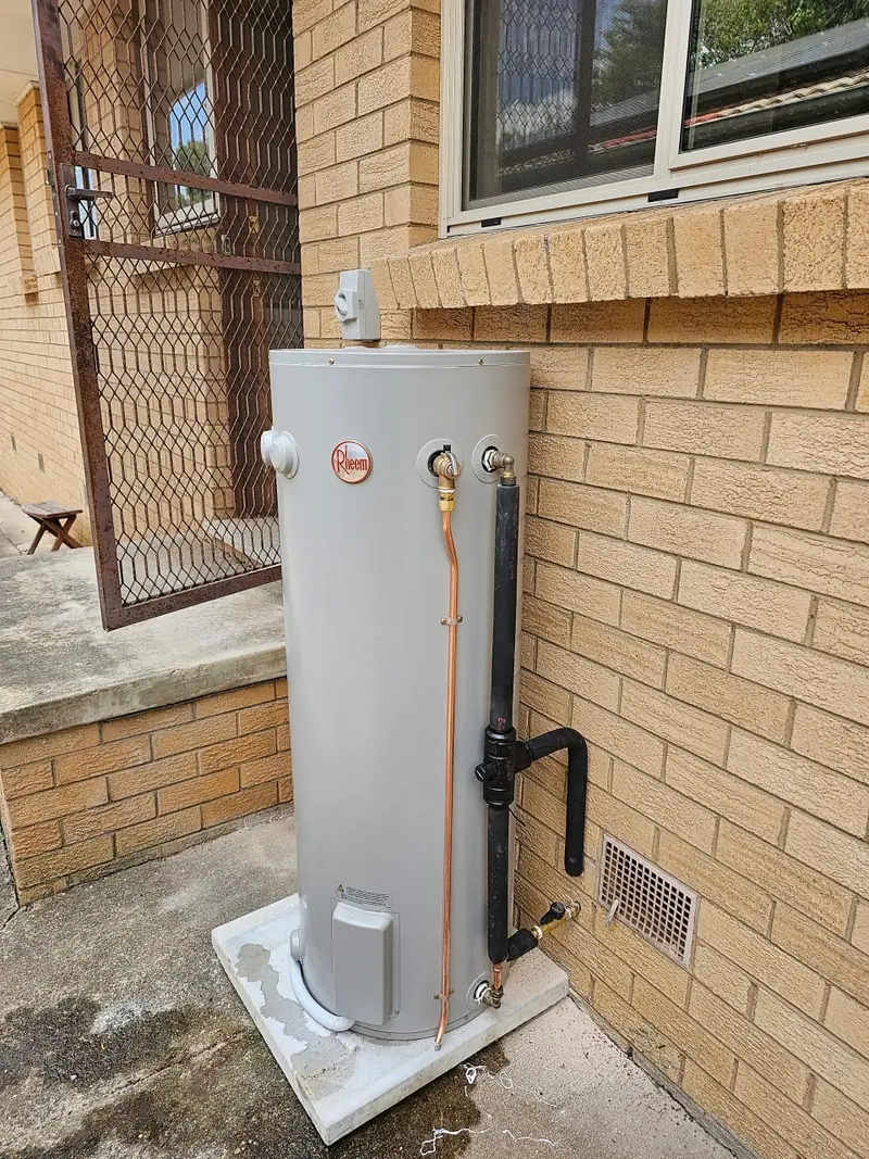An electric hot water cylinder installed on a concrete base against a brick wall, with new copper pipework run to it.